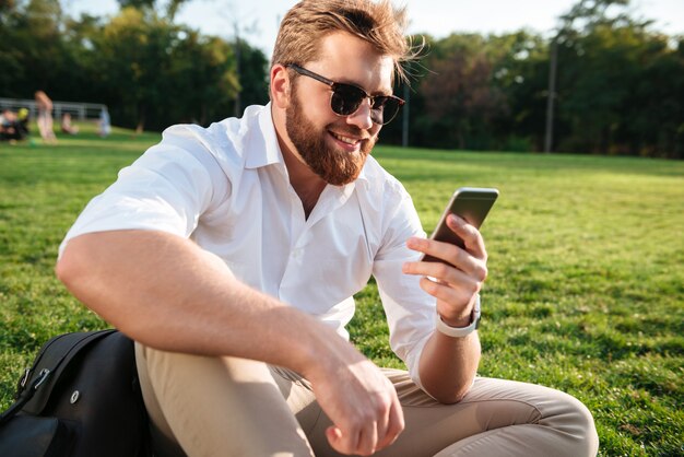 Hombre barbudo feliz en gafas de sol y ropa de negocios sentado en el césped al aire libre y usando su teléfono inteligente
