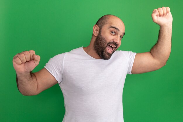 Hombre barbudo feliz loco en camiseta blanca sonriendo alegremente levantando los puños de pie sobre la pared verde