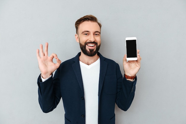 Hombre barbudo feliz en ropa de negocios que muestra la pantalla del teléfono inteligente en blanco