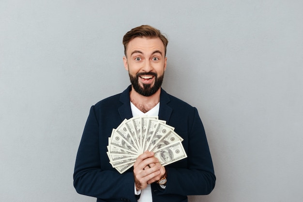 Hombre barbudo feliz sorprendido en ropa de negocios con dinero y mirando a la cámara sobre gris