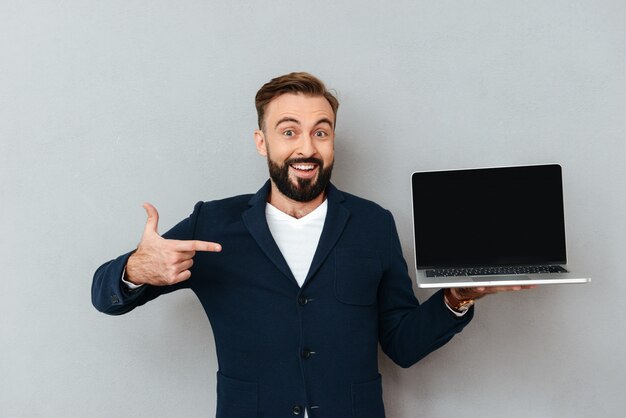 Hombre barbudo feliz sorprendido en ropa de negocios que muestra la pantalla de la computadora portátil en blanco y apuntando a él sobre gris