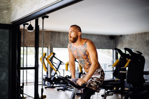 Hombre barbudo fuerte musculoso tatuado hacer cardio en bicicleta en el gimnasio cerca de una ventana grande con vista a los árboles afuera