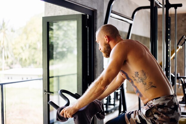 Hombre barbudo fuerte musculoso tatuado hacer cardio en bicicleta en el gimnasio cerca de una ventana grande con vista a los árboles afuera