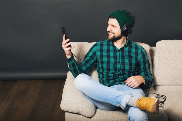 Hombre barbudo guapo joven inconformista sentado en un sofá en casa, escuchando música en auriculares, mirando smartphone, camisa a cuadros verde, entretenimiento