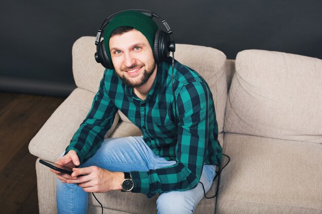 Hombre barbudo guapo joven inconformista sentado en un sofá en casa, escuchando música en auriculares