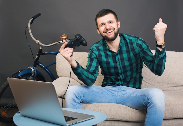 Foto gratuita hombre barbudo guapo joven inconformista sentado en el sofá en casa, jugando videojuegos en el portátil