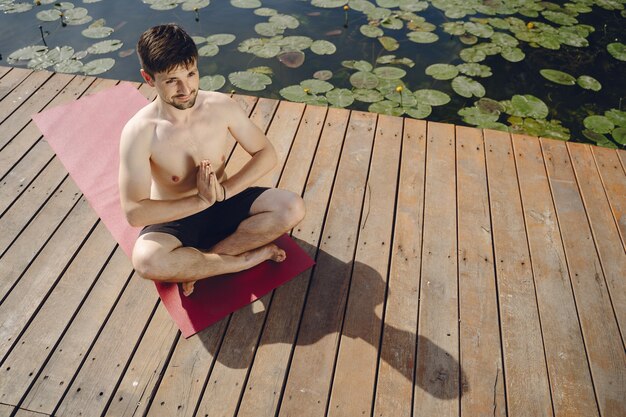 Hombre barbudo guapo joven sentado en el muelle de madera en día de verano. Meditar y relajarse.