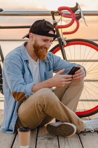 Foto gratuita hombre barbudo hipster guapo con smartphone viajando con bicicleta en el amanecer de la mañana junto al mar, viajero de estilo de vida activo y saludable