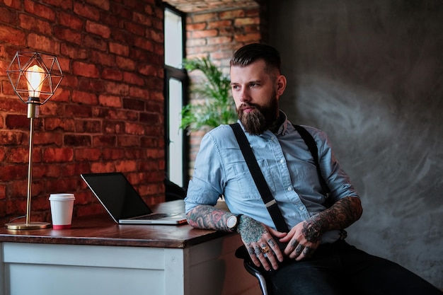 El hombre barbudo imponente se apoya en la mesa de su oficina. Lleva camisa y tirantes. Tiene tatuajes en los brazos. Hay una computadora portátil y una taza de café al fondo.