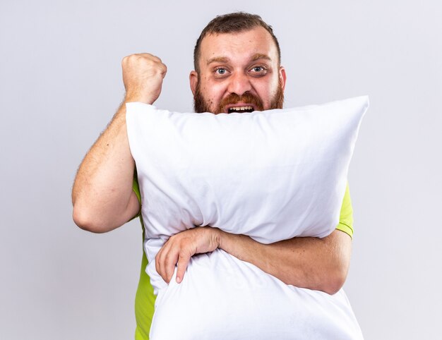 Hombre barbudo insalubre en camisa polo amarilla que se siente enfermo abrazando la almohada con la cara enojada levantando el puño sobre la pared blanca