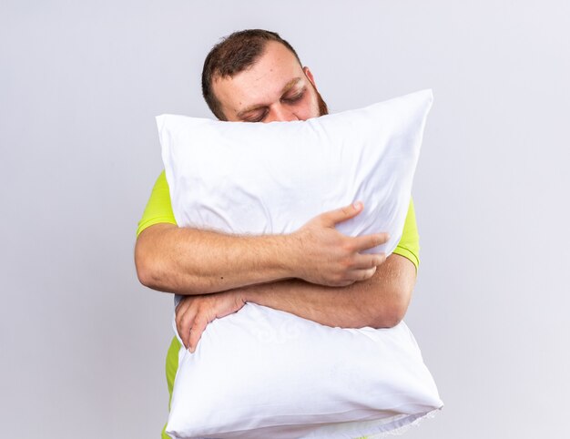 Hombre barbudo insalubre en camisa polo amarilla que se siente enfermo abrazando la almohada quiere dormir con los ojos cerrados expresión de pie sobre la pared blanca