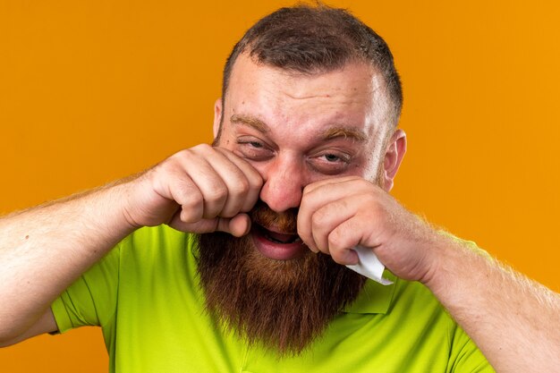 Hombre barbudo insalubre en camisa polo amarilla que se siente terrible que sufre de frío con dolor de cabeza debido a la nariz tapada de pie sobre la pared naranja