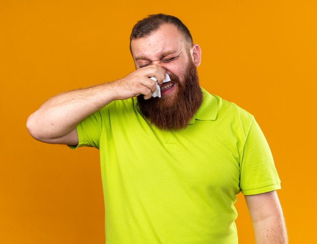 Hombre barbudo insalubre en camisa polo amarilla que se siente terrible sufriendo de frío limpiando su nariz que gotea