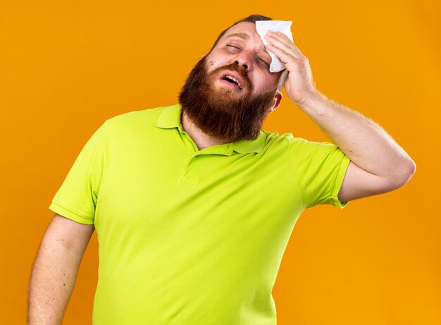 Hombre barbudo insalubre en camisa polo amarilla que se siente terrible sufrimiento de resfriado y fiebre sosteniendo tejido en la frente