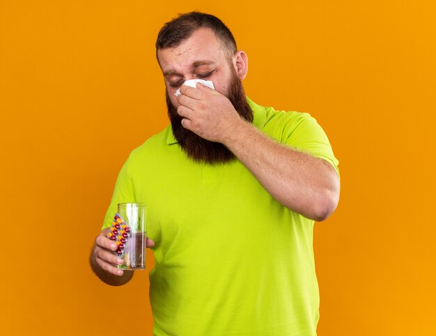 Hombre barbudo insalubre en camisa polo amarilla sosteniendo un vaso de agua y pastillas sintiéndose terrible sonarse la nariz en un tejido que sufre de frío parado sobre una pared naranja