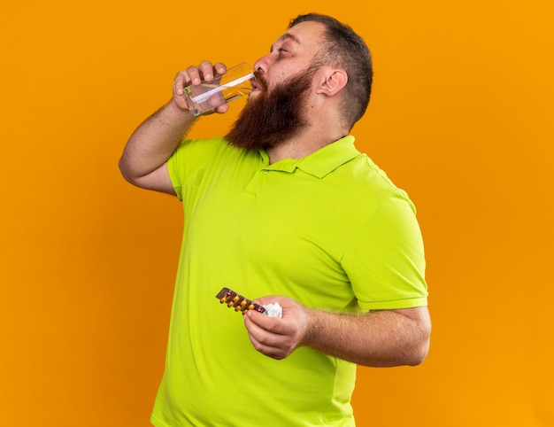 Hombre barbudo insalubre en camisa polo amarilla sosteniendo un vaso de agua y pastillas sintiéndose terrible sufrimiento de resfriado tomando medicamentos agua potable de pie sobre una pared naranja