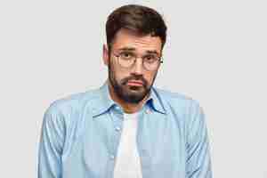 Foto gratuita el hombre barbudo insultado confundido se ve con expresión de duda miserable, tiene que tomar una decisión seria en la vida, usa gafas redondas y camisa azul, se para contra la pared blanca. concepto de personas y emociones