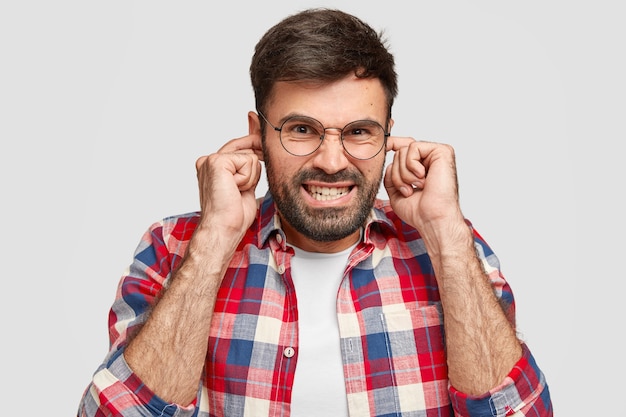 Hombre barbudo irritado tapona los oídos con molestia, escucha un sonido fuerte, aprieta los dientes, tiene expresión facial disgustada, usa una camisa a la moda, posa contra una pared blanca. ¡Detén este ruido!