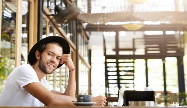 Hombre barbudo joven atractivo con sombrero negro de moda mirando con una sonrisa feliz, disfrutando de un buen café y un clima agradable mientras se relaja en el café solo durante el desayuno