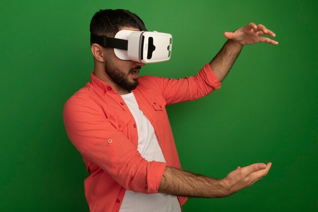 Hombre barbudo joven en camisa naranja con gafas de realidad virtual que muestran gesto de tamaño con las manos de pie sobre la pared verde