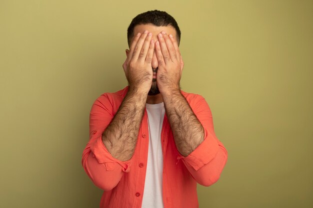 Hombre barbudo joven en camisa naranja que cubre los ojos con las manos de pie sobre la pared de luz