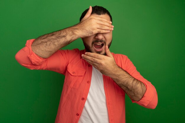 Hombre barbudo joven en camisa naranja que cubre su rostro con las manos sonriendo de pie sobre la pared verde