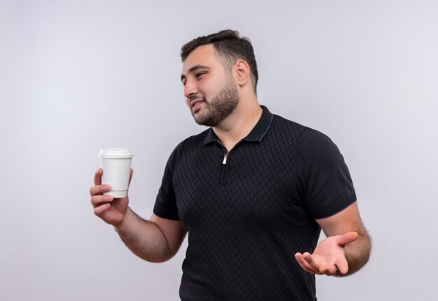 Hombre barbudo joven en camisa negra sosteniendo la taza de café mirando a un lado gesticulando con la mano