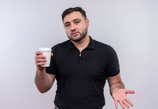 Hombre barbudo joven en camisa negra con taza de café mirando confundido