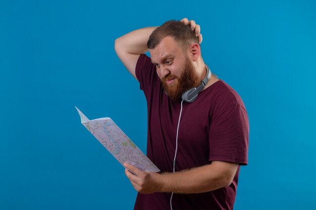 Hombre barbudo joven en camiseta marrón con auriculares alrededor de su cuello sosteniendo el mapa mirándolo desconcertado