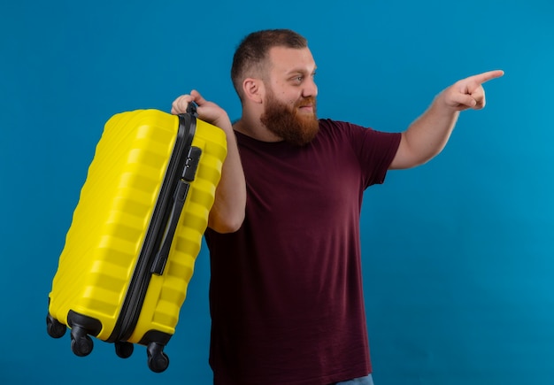 Hombre barbudo joven en camiseta marrón con maleta de viaje mirando a un lado apuntando con el dedo índice a algo positivo y feliz