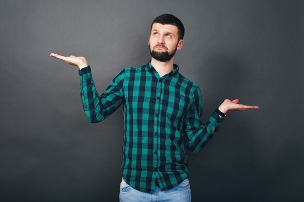 Hombre barbudo joven guapo hipster sosteniendo las manos arriba pregunta de evaluación, camisa a cuadros verde, fondo gris