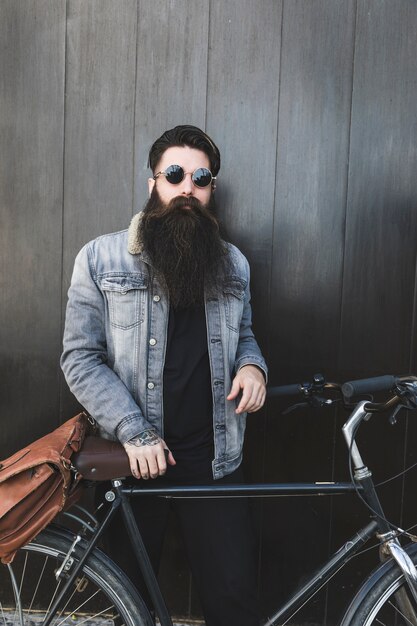 Hombre barbudo joven de moda que se coloca con la bicicleta delante de la pared de madera negra