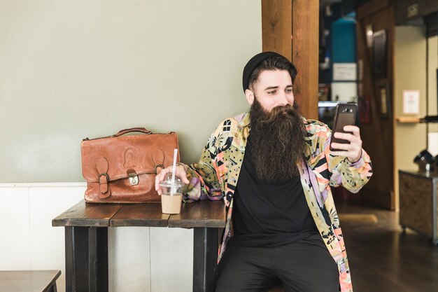 Hombre barbudo joven que se sienta en el café que toma el selfie en el teléfono móvil