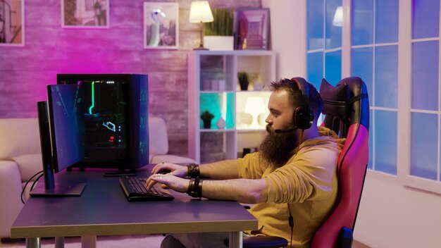 Hombre barbudo jugando videojuegos en una habitación con neones de colores. Hombre hablando con sus amigos mientras juega videojuegos.