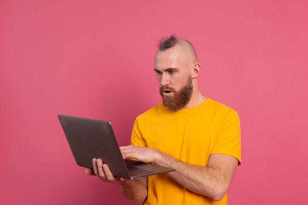 Hombre barbudo con laptop emociones de choque aislado sobre fondo rosa