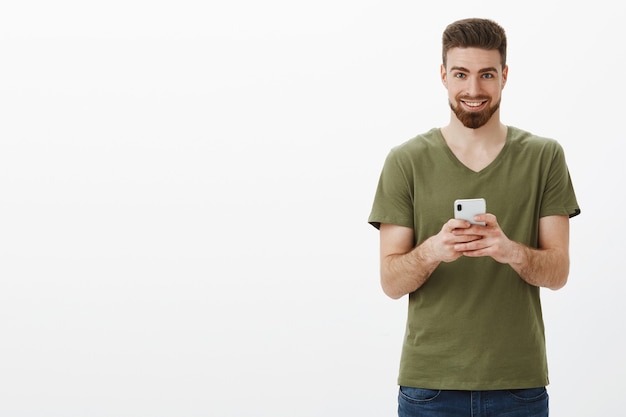 Hombre barbudo maduro, guapo, retorcido y emocionado, con camiseta verde oliva, sosteniendo un teléfono inteligente y luciendo complacido sonriendo como si tuviera una idea interesante de la próxima publicación en línea sobre una pared blanca