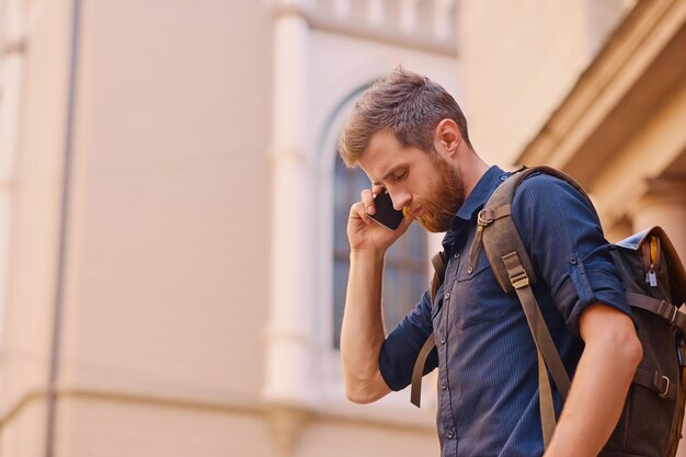 Hombre barbudo con mochila hablando por teléfono inteligente en una antigua ciudad europea.