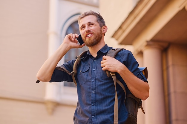 Hombre barbudo con mochila hablando por teléfono inteligente en una antigua ciudad europea.