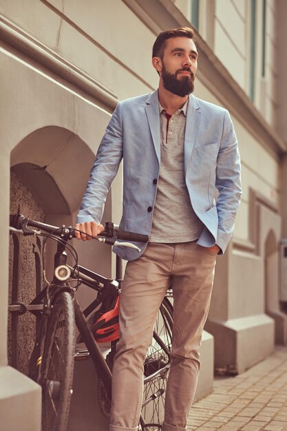 Hombre barbudo de moda con ropa elegante, se para con una bicicleta en una calle antigua en Europa.
