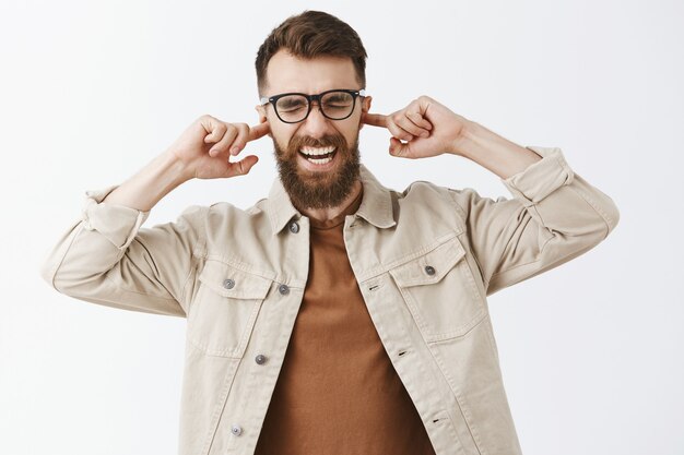 Hombre barbudo molesto enojado con gafas posando contra la pared blanca