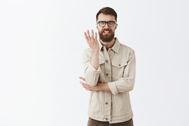 Hombre barbudo molesto y frustrado con gafas posando contra la pared blanca