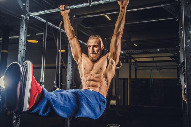 Hombre barbudo musculoso sin camisa haciendo ejercicios en la barra horizontal en un club de gimnasia.