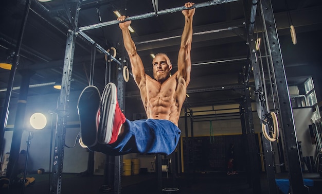 Hombre barbudo musculoso sin camisa haciendo ejercicios en la barra horizontal en un club de gimnasia.