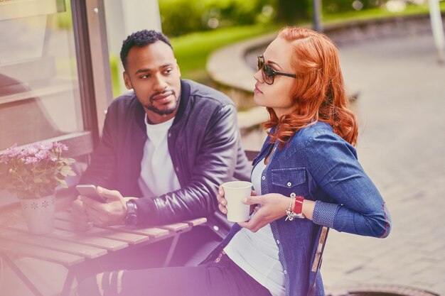 Hombre barbudo negro y mujer pelirroja beben café en una cafetería en una calle.