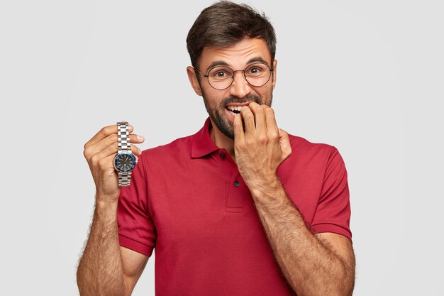 Hombre barbudo nervioso se muerde las uñas, sostiene el reloj de pulsera, se preocupa por llegar tarde a una reunión importante, vestido con una camiseta informal. Joven avergonzado espera algo. El tiempo vuela rápido