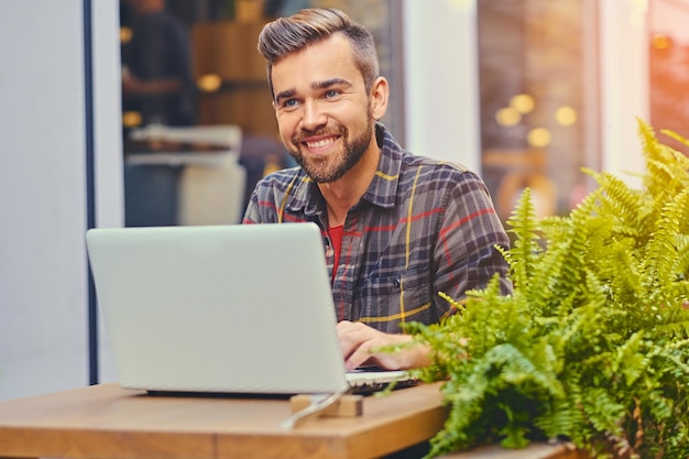 Hombre barbudo de ojos azules que usa una computadora portátil en un café en una calle.