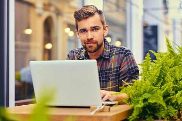 Hombre barbudo de ojos azules que usa una computadora portátil en un café en una calle.