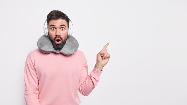 Hombre barbudo sin palabras aturdido tiene una barba gruesa en el espacio de la copia que promueve algo increíble usa una almohada cómoda alrededor del cuello usa audífonos para escuchar música