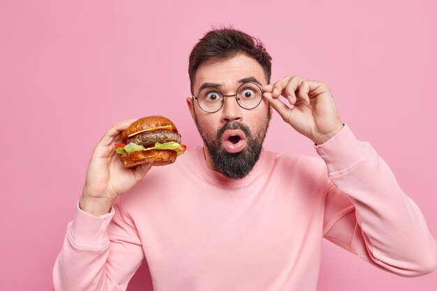 Hombre barbudo sin palabras, sorprendido, mantiene la mano en el borde de las gafas, descubre noticias impactantes sobre lo dañina que es la comida rápida, sostiene apetitosa hamburguesa, usa jersey informal