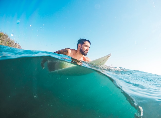 Hombre barbudo en pantalones cortos sentado en tabla de surf en agua